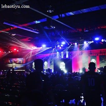 A dimly lit indoor arena filled with an audience. Bright stage lights in red and blue illuminate multiple large screens displaying graphics.