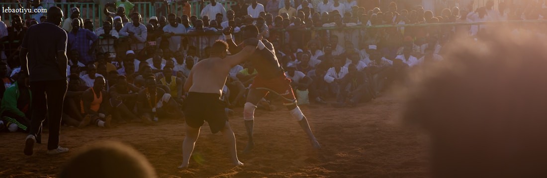 A vibrant wrestling match taking place outdoors, surrounded by a large crowd of spectators. The atmosphere is charged with excitement as people stand and sit around the dirt arena, intently watching the competitors. The sun is setting, casting a warm glow over the scene.