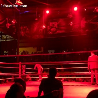 A wrestling ring is illuminated with red lighting, creating a dramatic atmosphere. Several people are visible around the ring, with silhouetted spectators in the foreground. Banners or art adorn the upper walls, and the venue has an industrial ceiling with exposed beams.